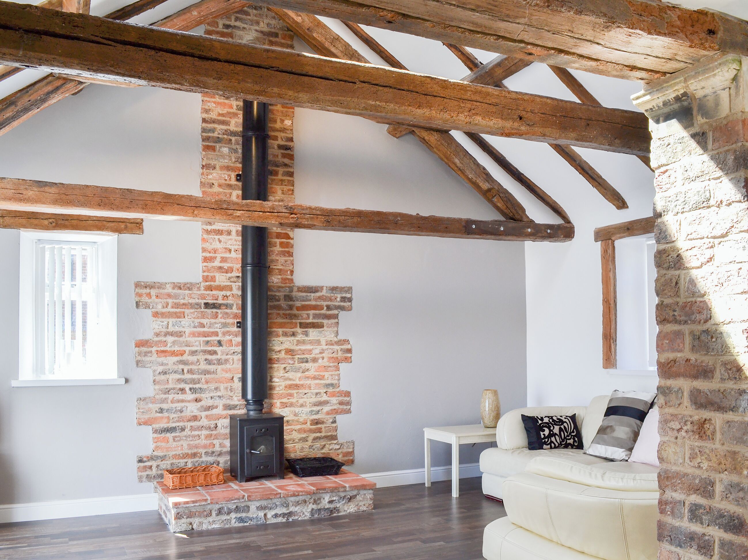 A room with exposed wooden beams, a brick fireplace, a white sofa, and a small table with a vase.