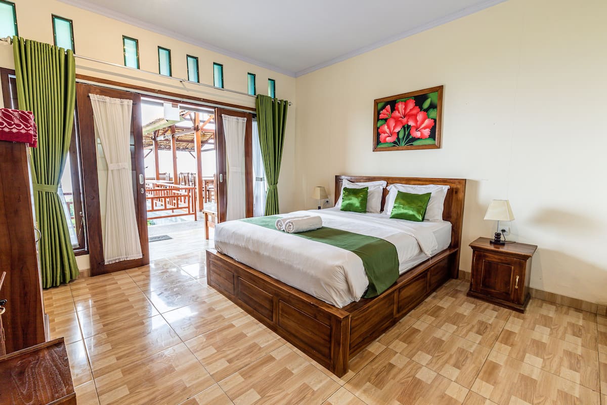 A bedroom with a wooden bed, a nightstand, a painting of flowers, and a view of an outdoor area.