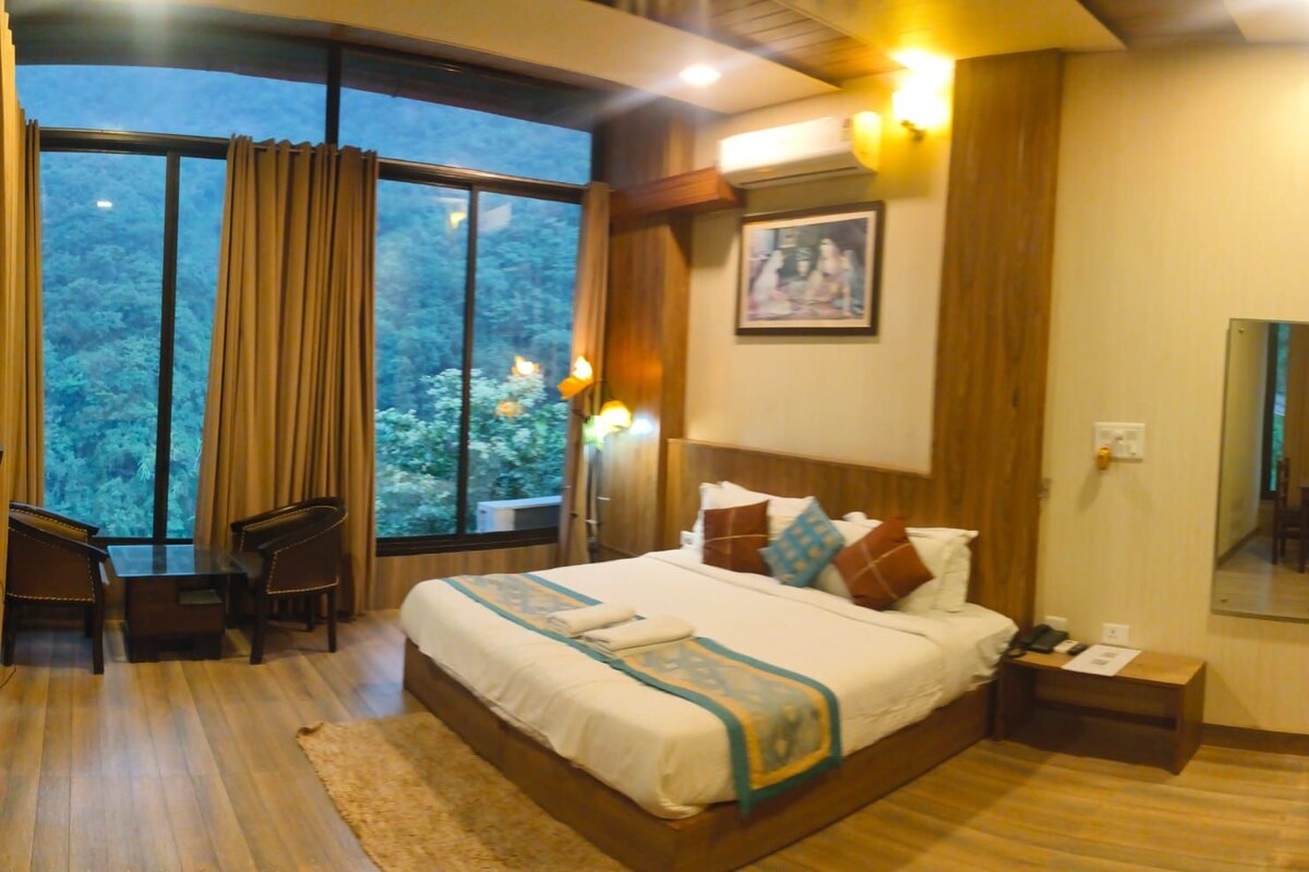 A hotel room with a large bed, a bedside table, a chair, a window with curtains, and a view of trees.