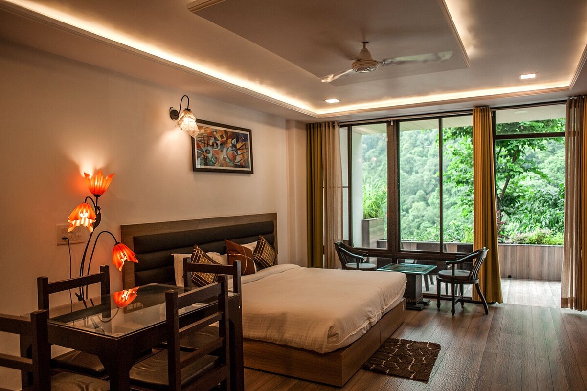 A modern hotel room with a large bed, a desk, and a view of greenery through the windows.