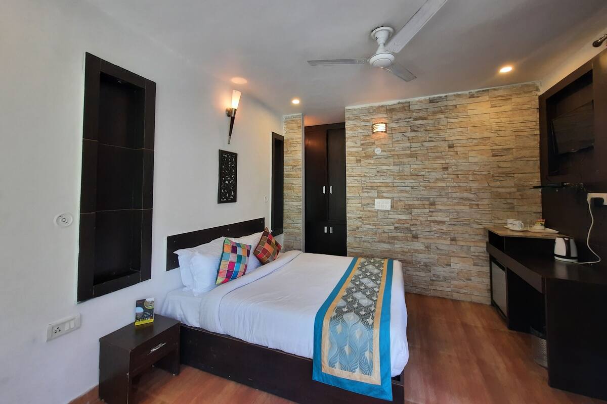 A bedroom with a bed, a nightstand, a bedside table, a ceiling fan, a stone wall, a TV, and a small table with a cup and saucer.