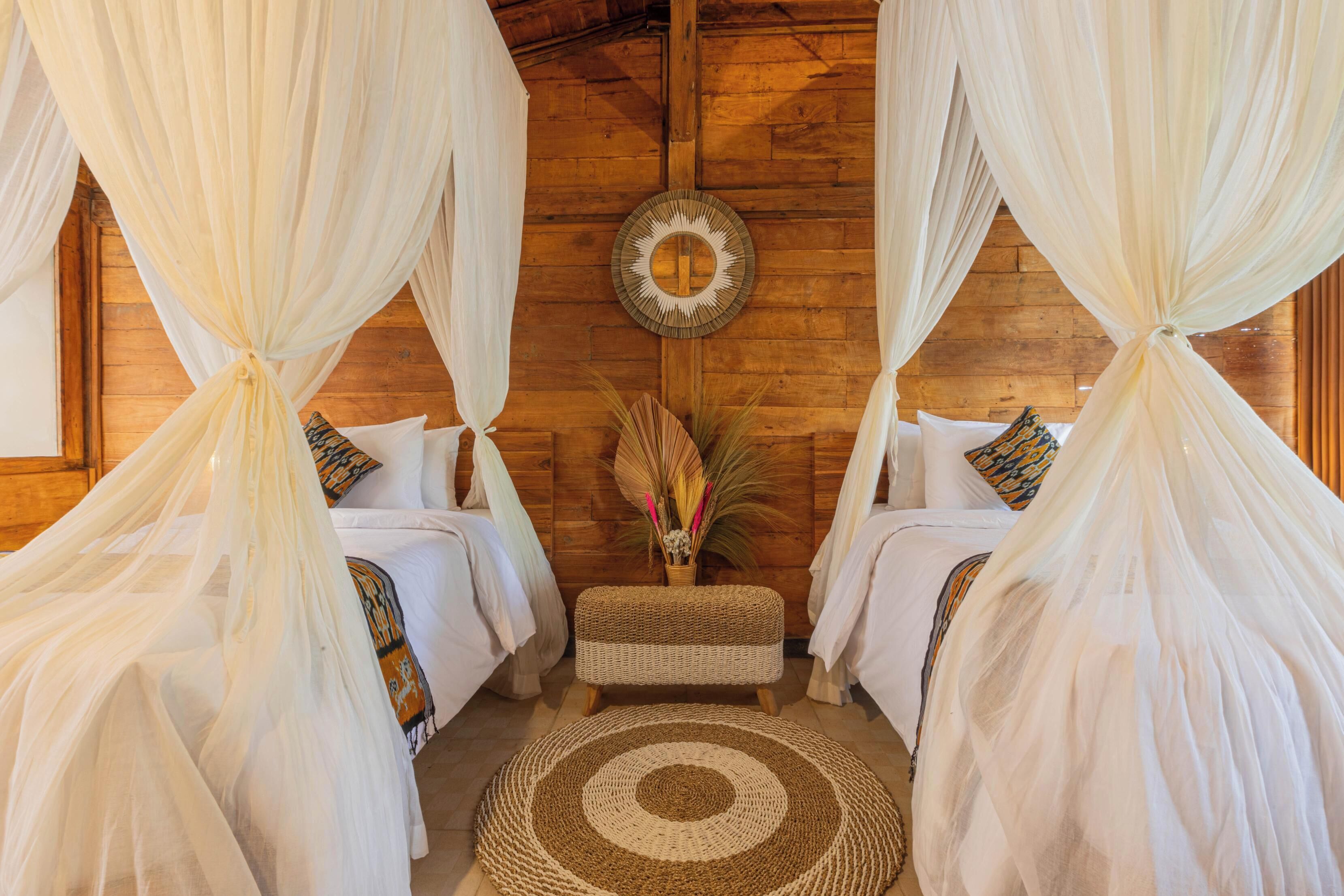 A room with two beds, each with white bedding and a canopy. There is a woven bench between the beds, and a round woven rug on the floor.