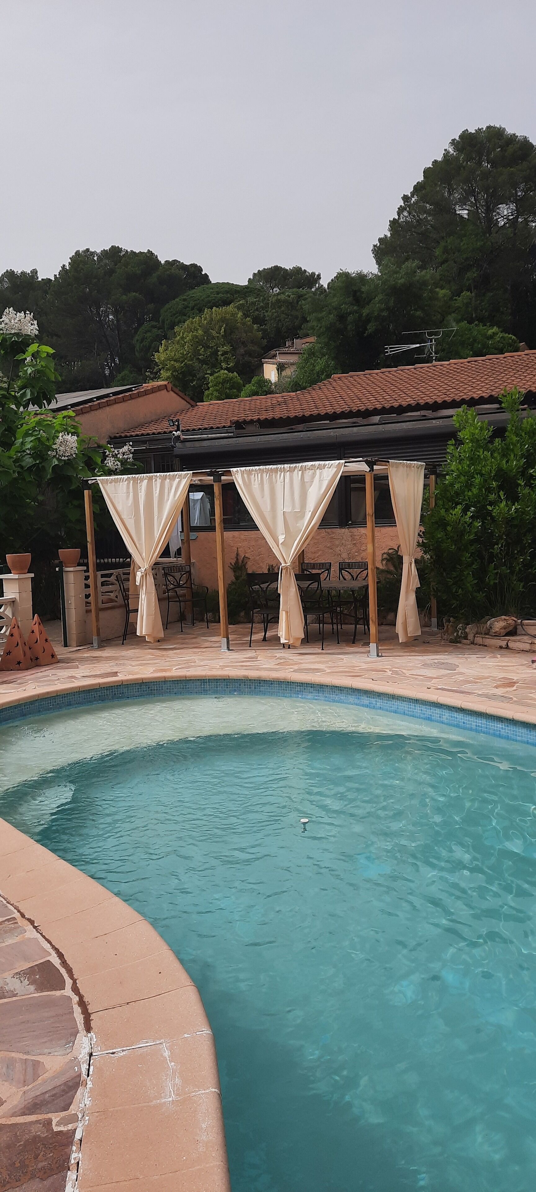 Une piscine avec un bord carrelé, un espace détente couvert avec des chaises, et une pergola agrémentée de rideaux.