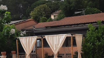 Une piscine avec un bord carrelé, un espace détente couvert avec des chaises, et une pergola agrémentée de rideaux.