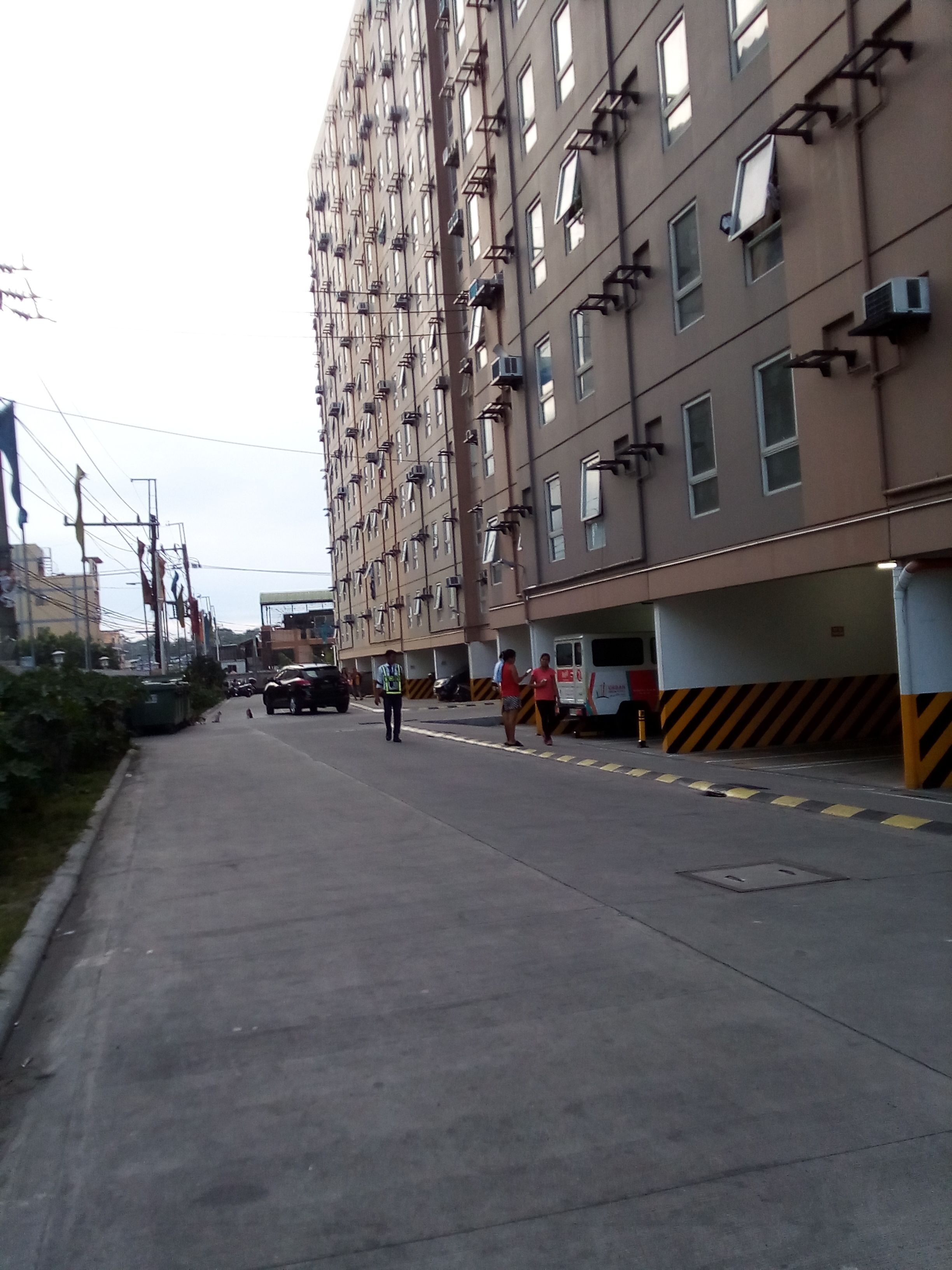 A multi-story residential building with balconies, a parking area, and a sidewalk.