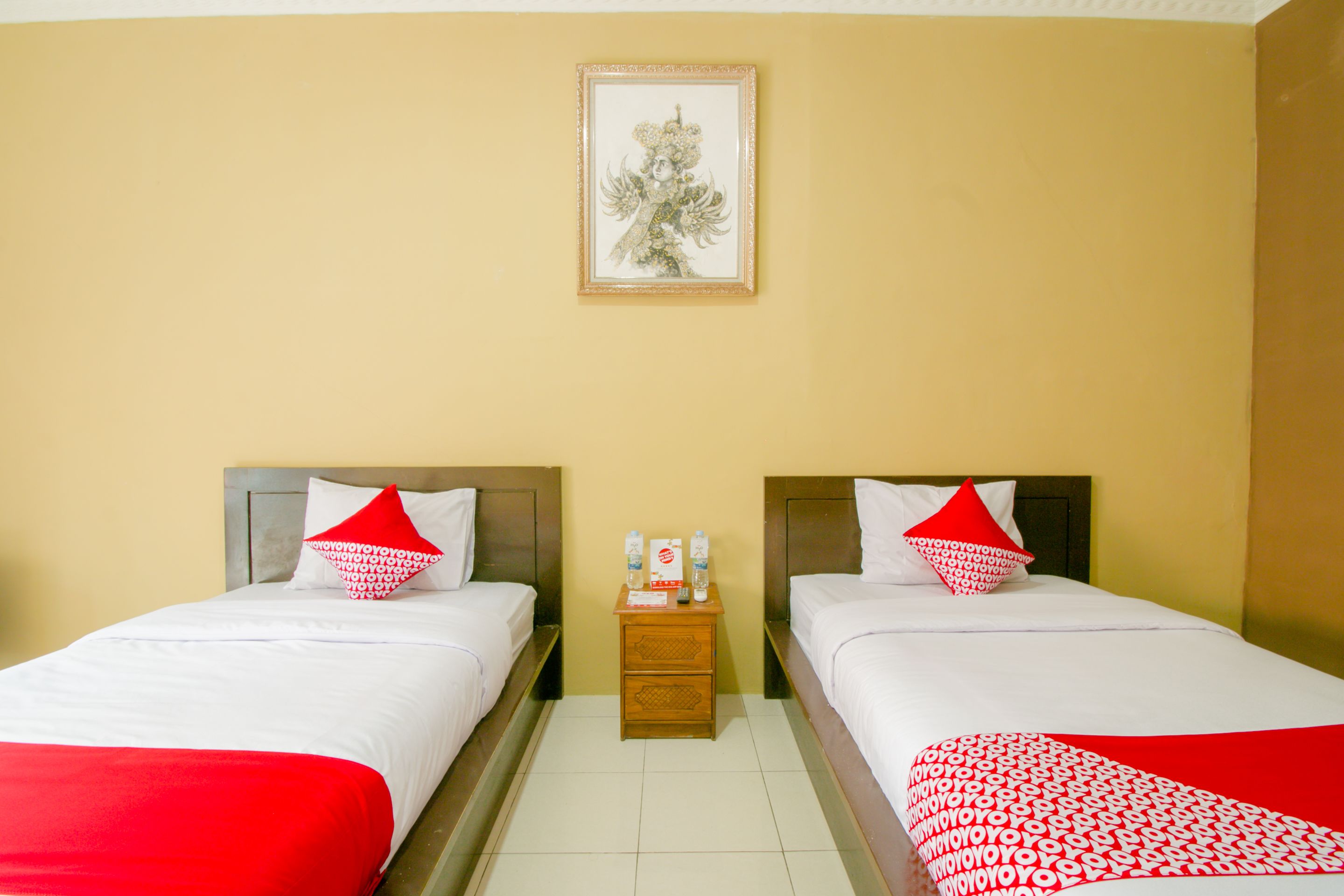 Hotelkamer met twee bedden, elk voorzien van witte beddengoed en rode accenten, een klein houten nachtkastje met toiletartikelen, en een lijst met kunstwerk aan de muur.
