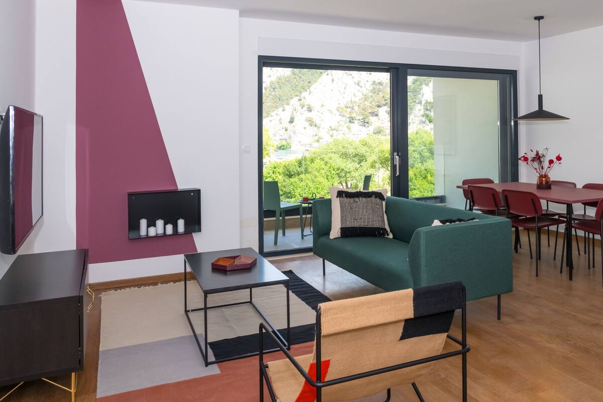 Un salon moderne avec un canapé vert, un coin repas avec des chaises rouges et une grande fenêtre donnant sur un paysage verdoyant.