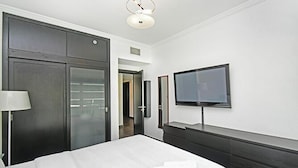 A modern hotel room with a large bed, a flat-screen TV mounted on the wall, a dark wooden dresser, and a walk-in closet. - Al Majara 5 by Deluxe Holiday Homes (Dubai)
