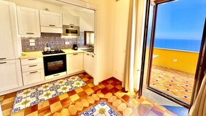 Una cucina con mobili bianchi, un fornello e vista sul mare tramite una porta scorrevole in vetro.