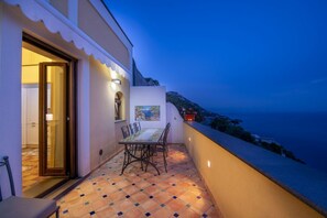 A balcony with a table and chairs, a painting, and a view of the sea at dusk.