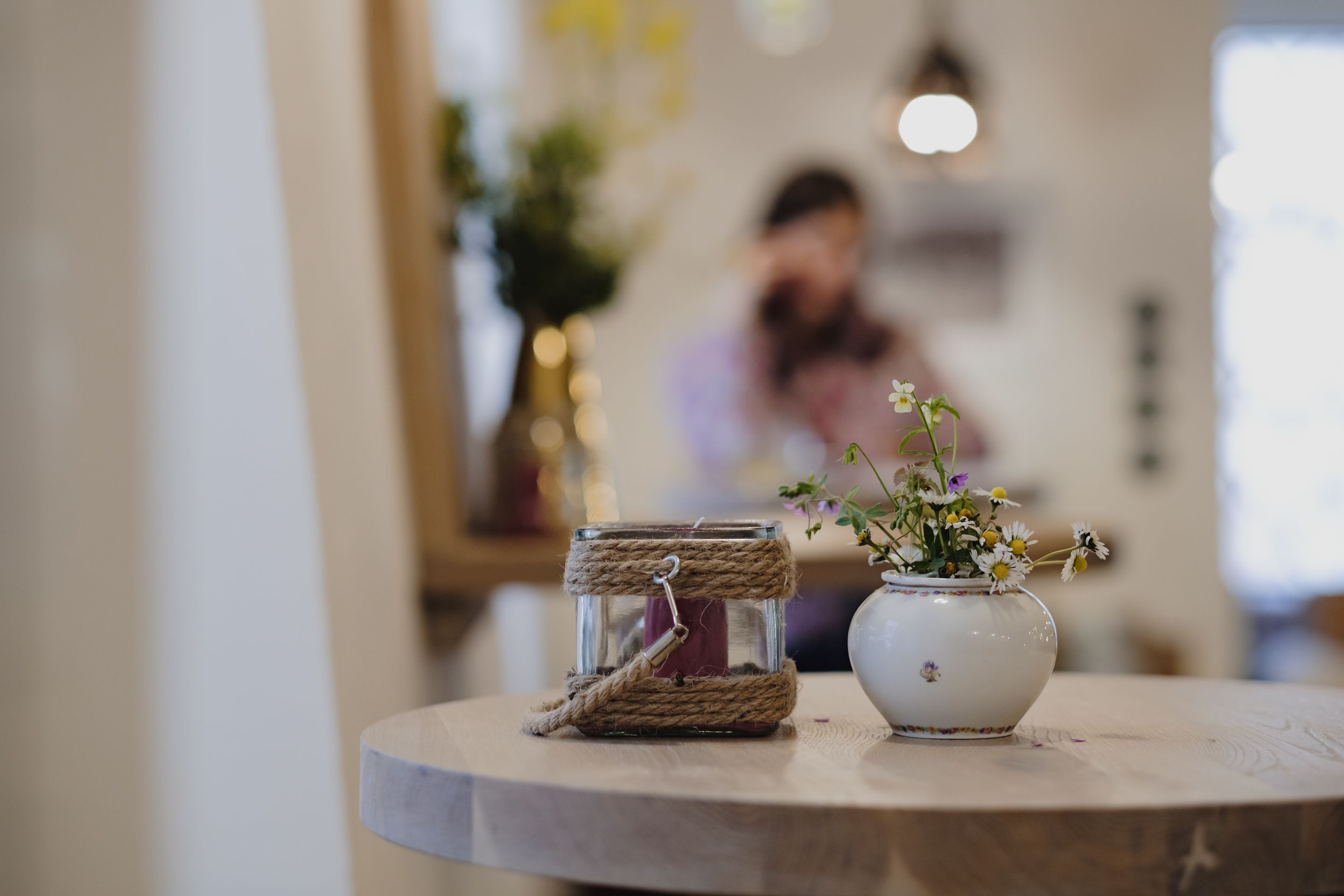 A round wooden table with a glass jar wrapped in twine and a white ceramic vase with flowers.