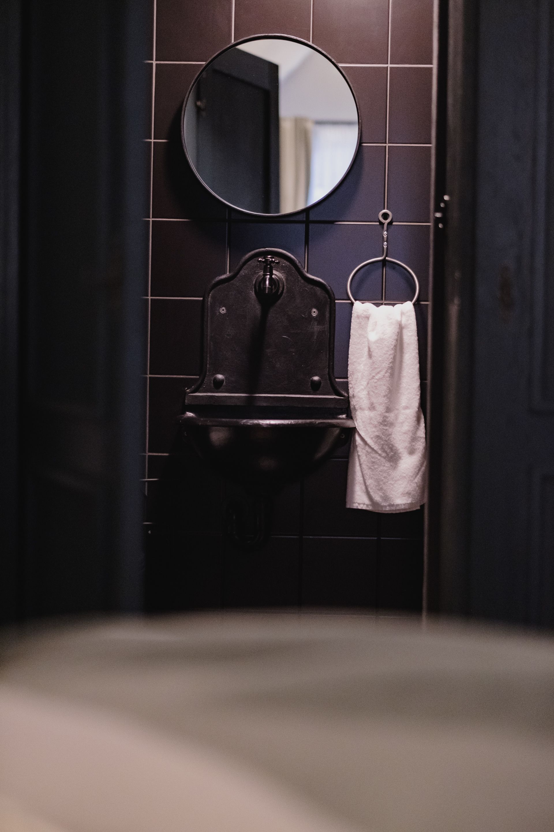A bathroom with a dark tiled wall, a round mirror, a sink, and a towel hanging on a rack.