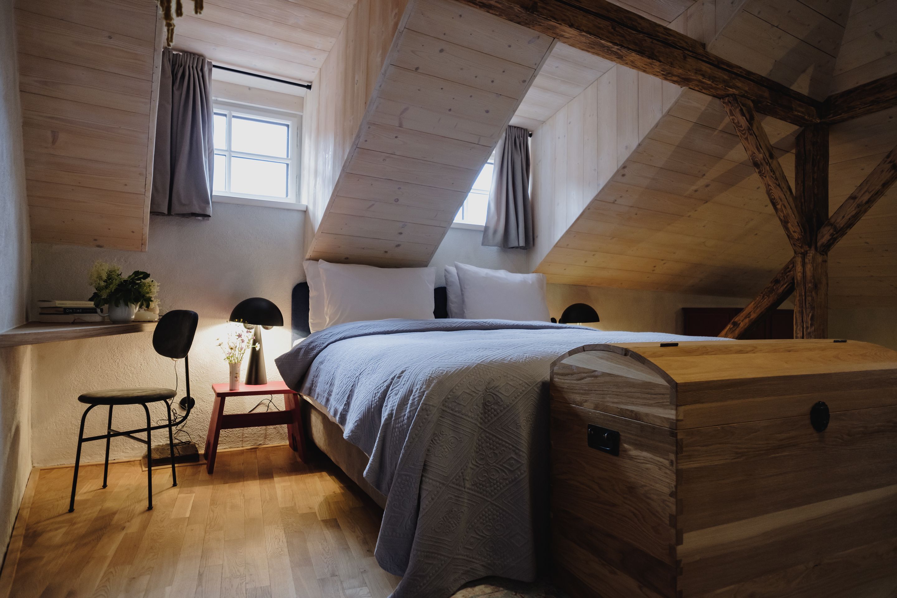 Un dormitorio acogedor en el ático con techo de madera, una cama con colcha gris, una mesita de noche con lámpara y una silla.