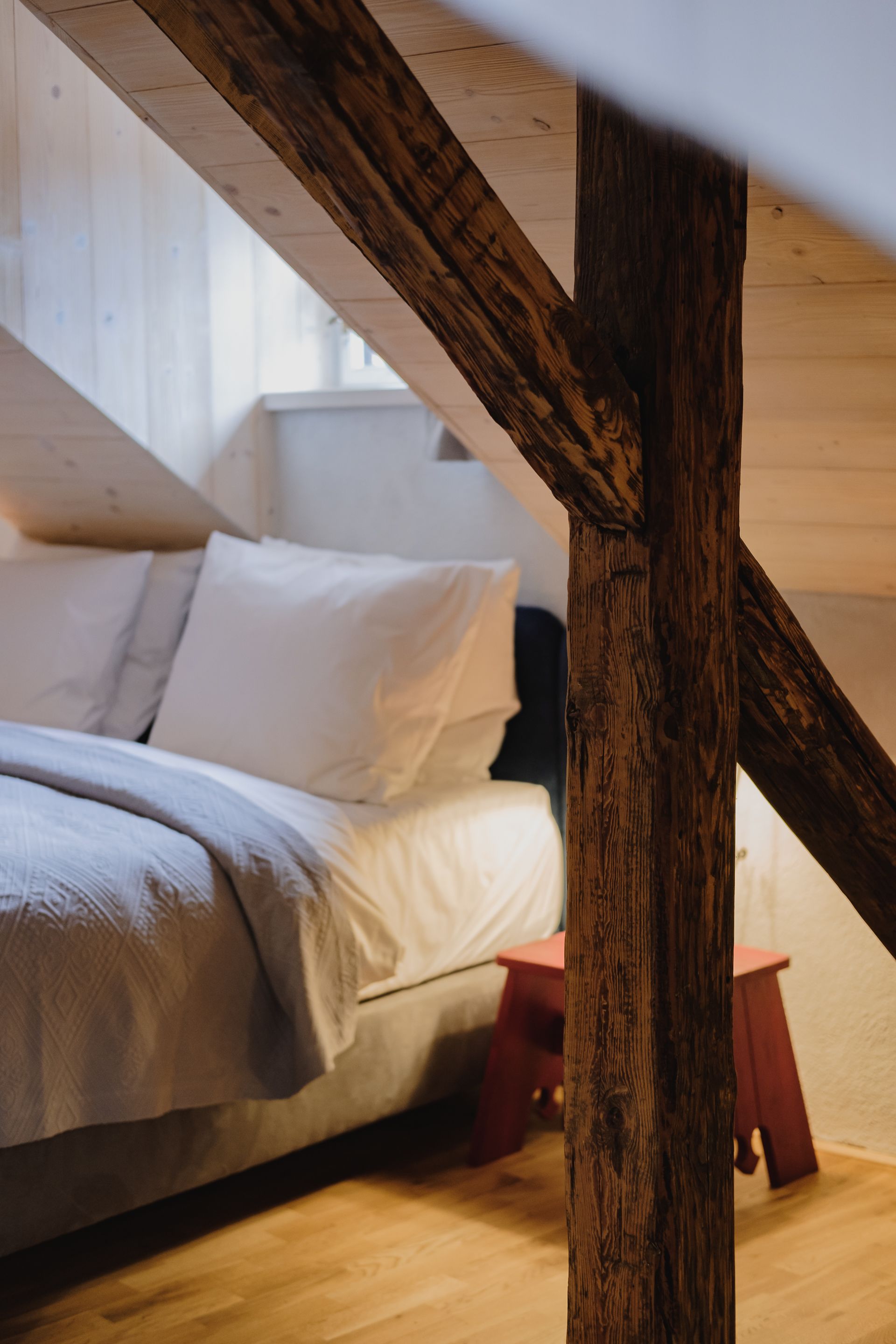 A bedroom with a sloped ceiling, wooden beams, a bed with white bedding, and a small red stool.