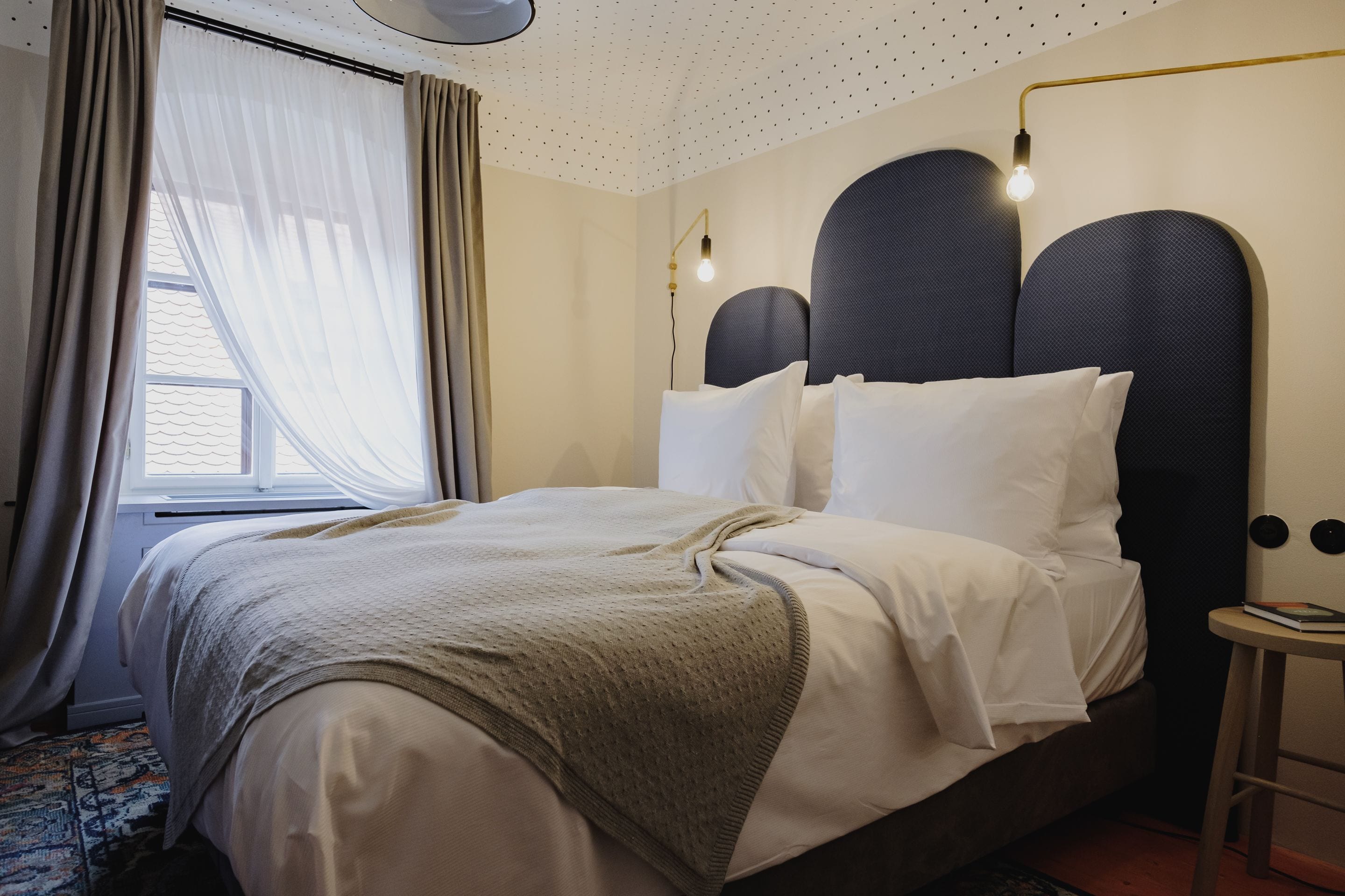 A neatly made bed with a headboard, a bedside table, and a window with curtains.