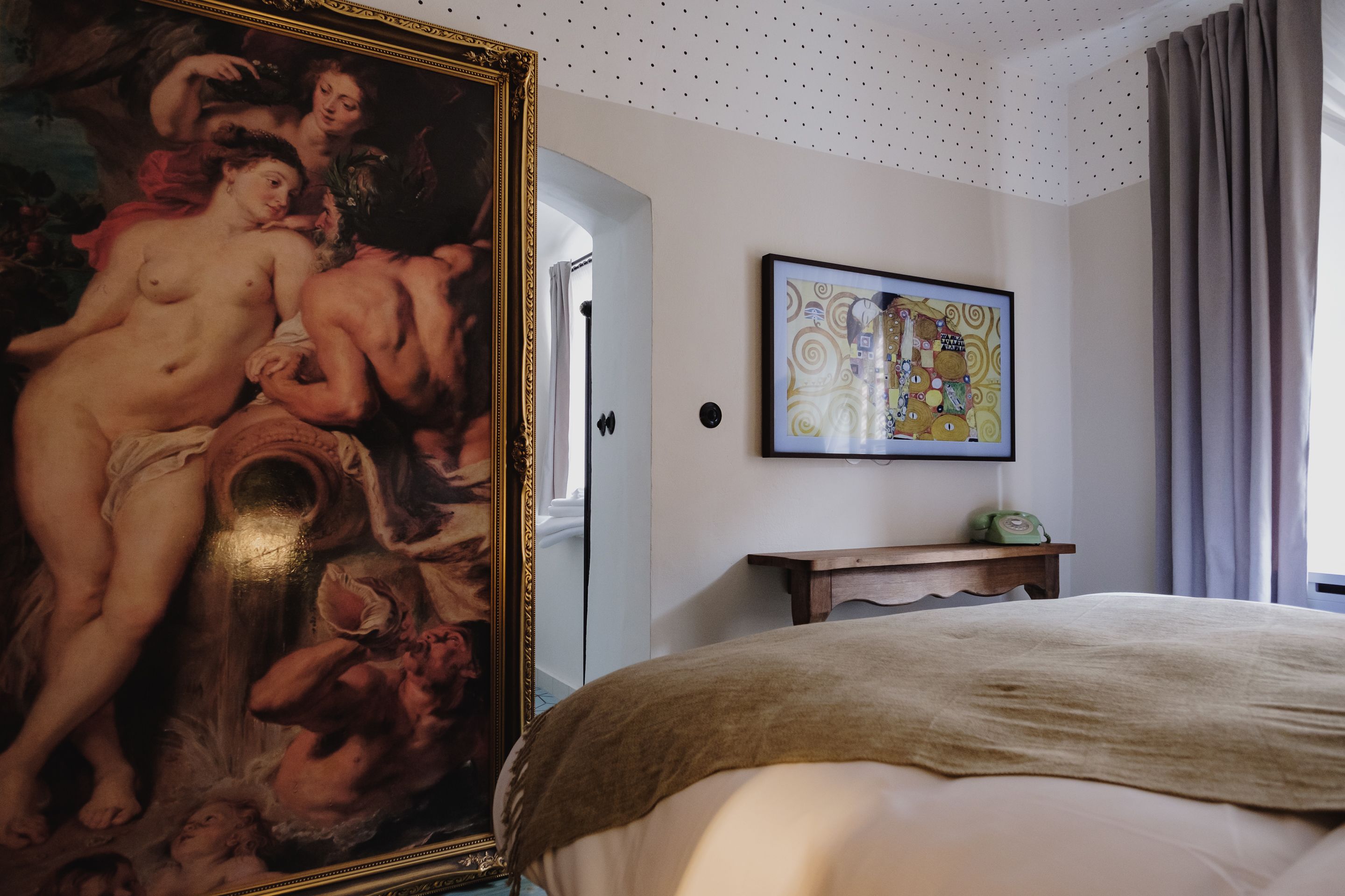 A bedroom with a large classical painting, a modern abstract painting, and a bed with a beige blanket.
