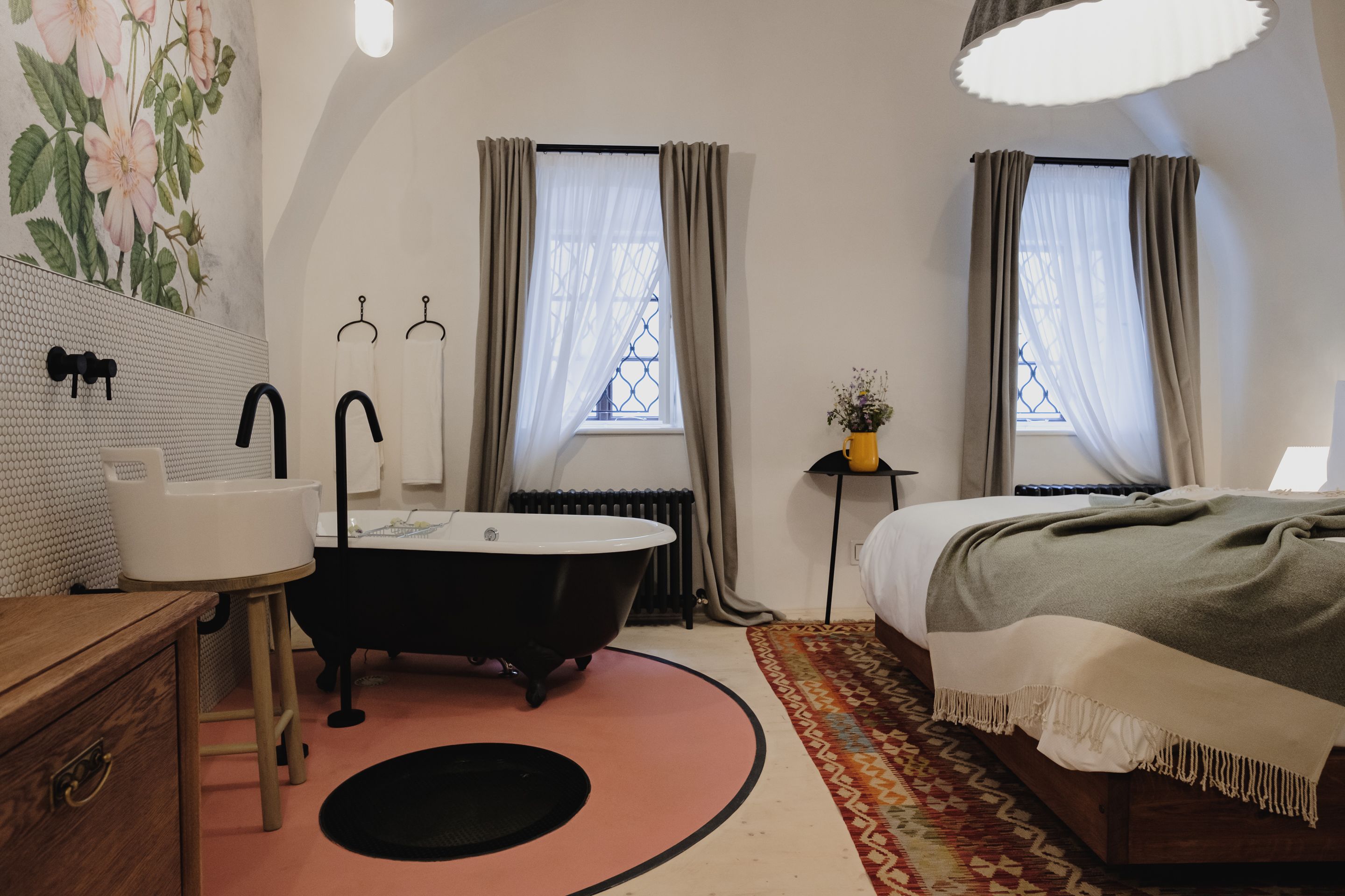A bedroom with a bed, a bedside table, a nightstand, a window with curtains, a radiator, and a bathroom with a bathtub, sink, and mirror.