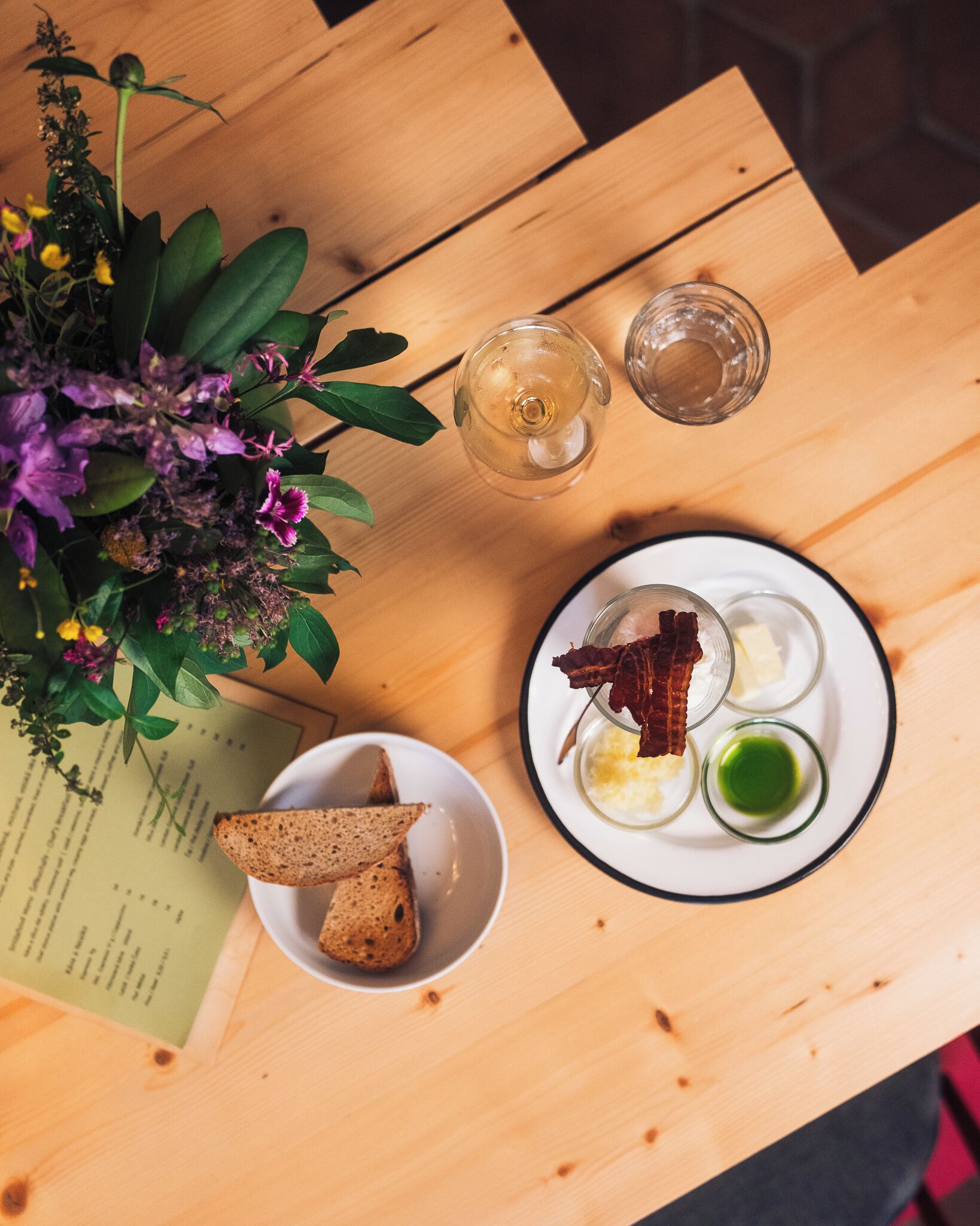 Una mesa de madera con un menú, un plato de comida que incluye tostadas y un bol pequeño, y una copa de vino.