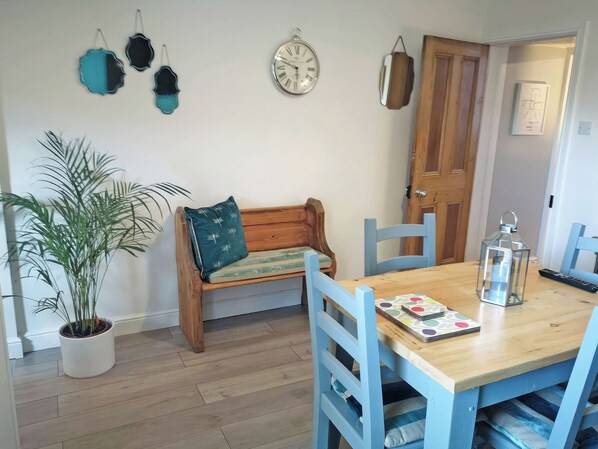 A dining area with a wooden table, blue chairs, a bench with a cushion, a potted plant, wall-mounted clocks, and a door.
