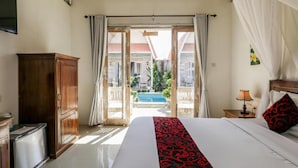 A hotel room with a bed, a desk, a TV, and a view of a pool and buildings. - The Box Paradise Bungalow (Penida Island)