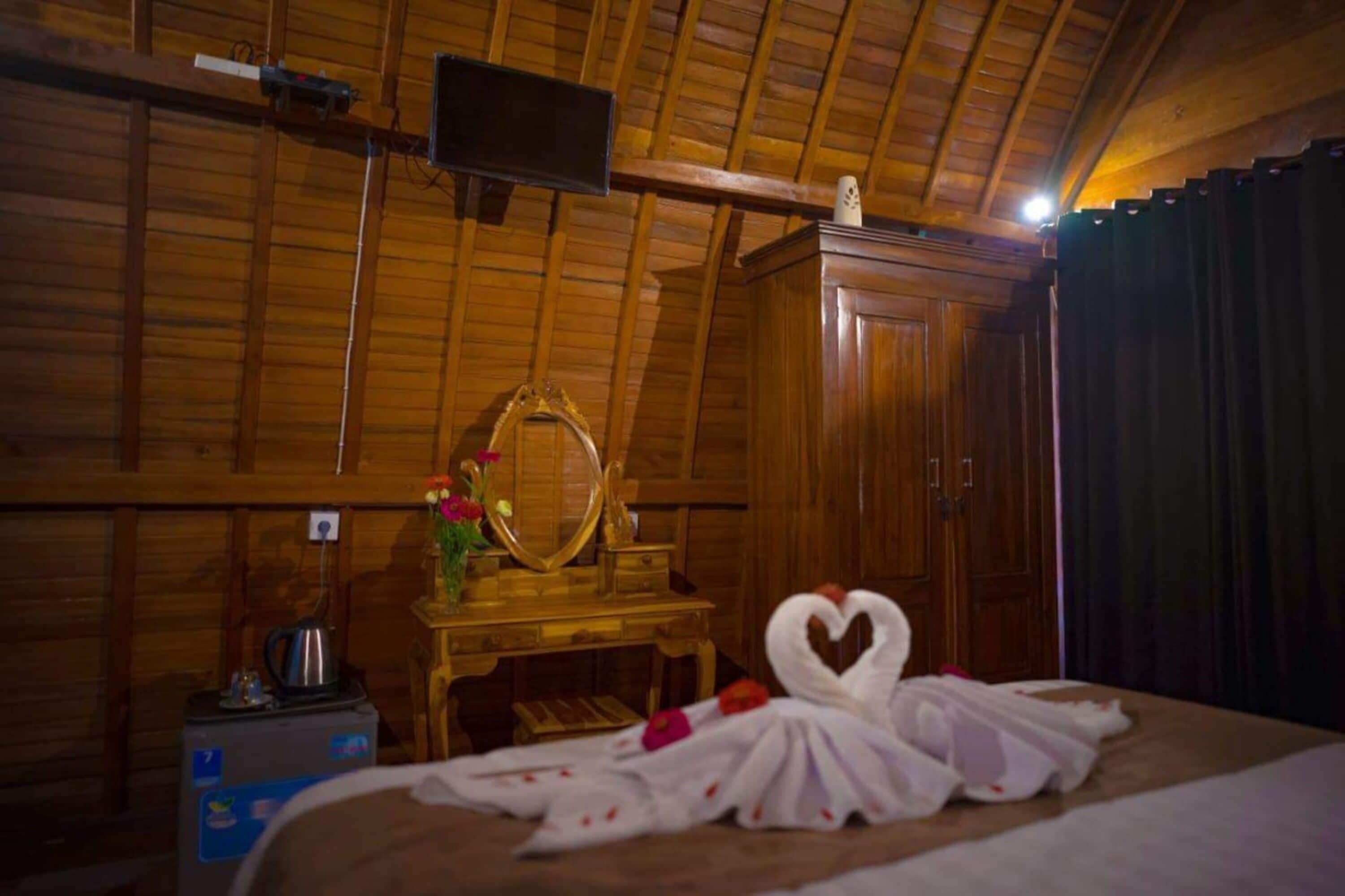 Une pièce en bois avec un lit, une armoire en bois, un miroir et une petite table sur laquelle se trouve un vase avec des fleurs.
