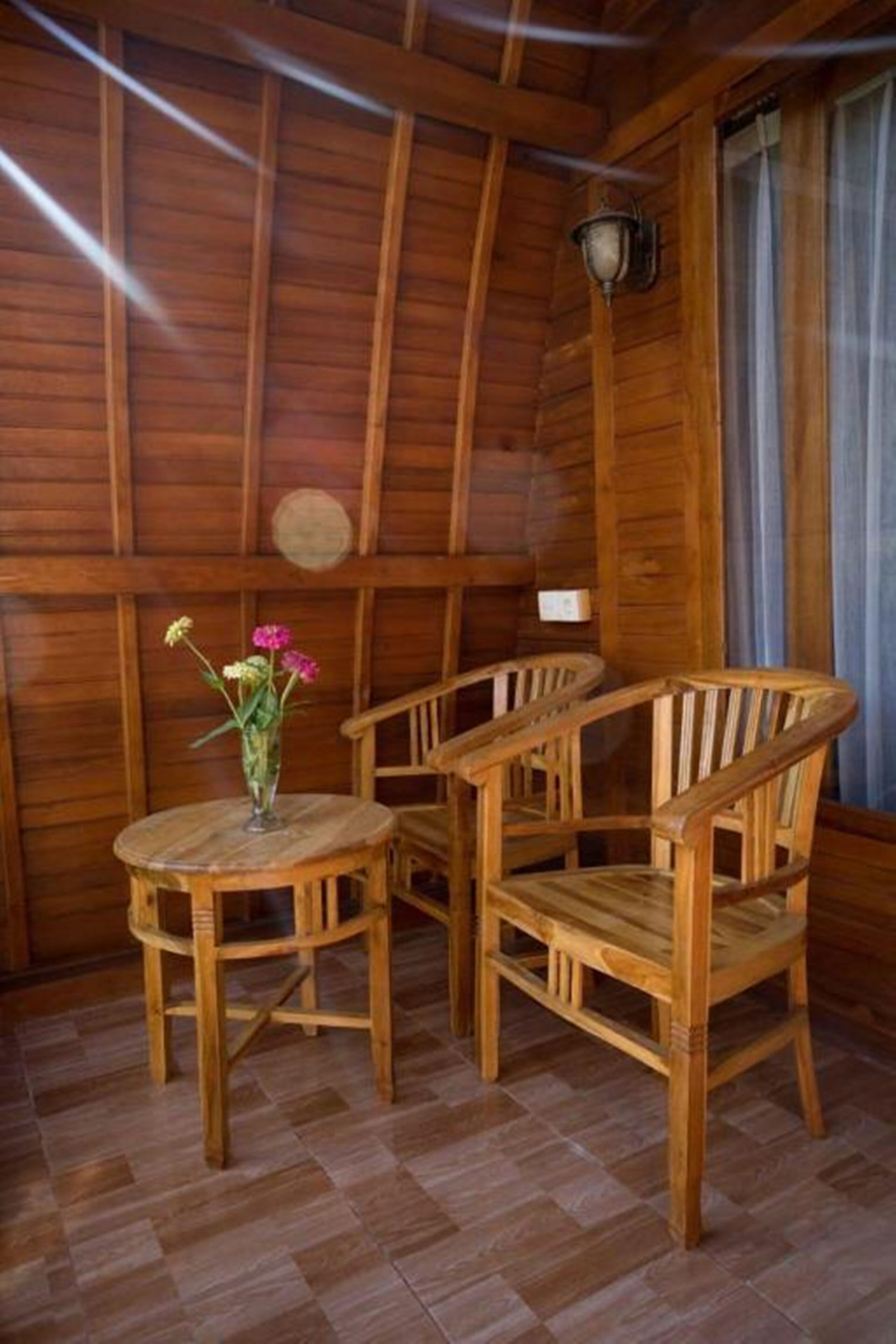 Een houten interieur met een ronde houten tafel en twee stoelen, een vaas met bloemen, en een wandlamp.