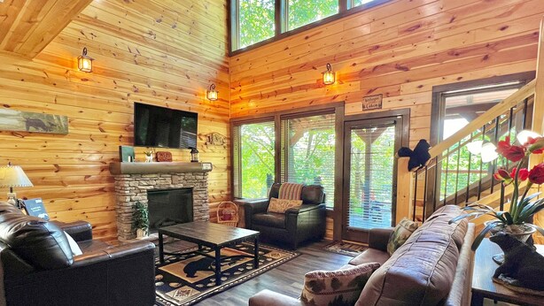 A cozy cabin interior with a stone fireplace, leather sofas, and a large window offering a view of the outdoors.