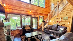 A cozy cabin interior with a stone fireplace, leather sofas, a staircase, and large windows offering a view of greenery.