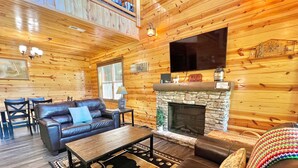 A cozy cabin interior with a stone fireplace, leather sofas, a dining area, and a large TV mounted on the wall.