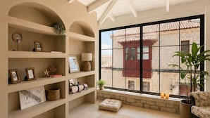 A spacious living room with high ceilings, a large window with a view of a stone building, and a cozy seating area with cushions and a rug. -  Als Alaçatı (Cesme)
