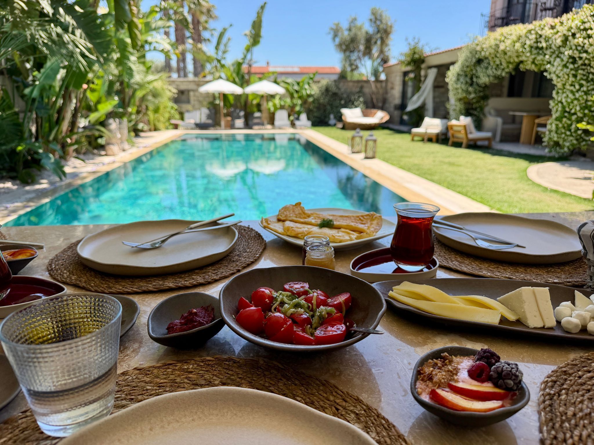 A dining table with plates, cutlery, and food items, overlooking a swimming pool and garden.