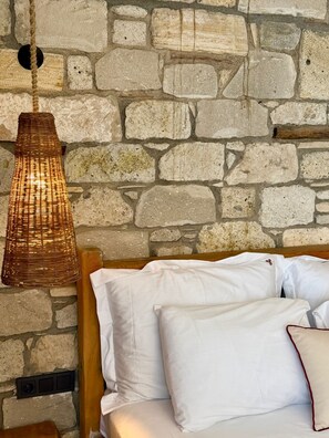 A bed with white bedding and a woven pendant light against a stone wall.