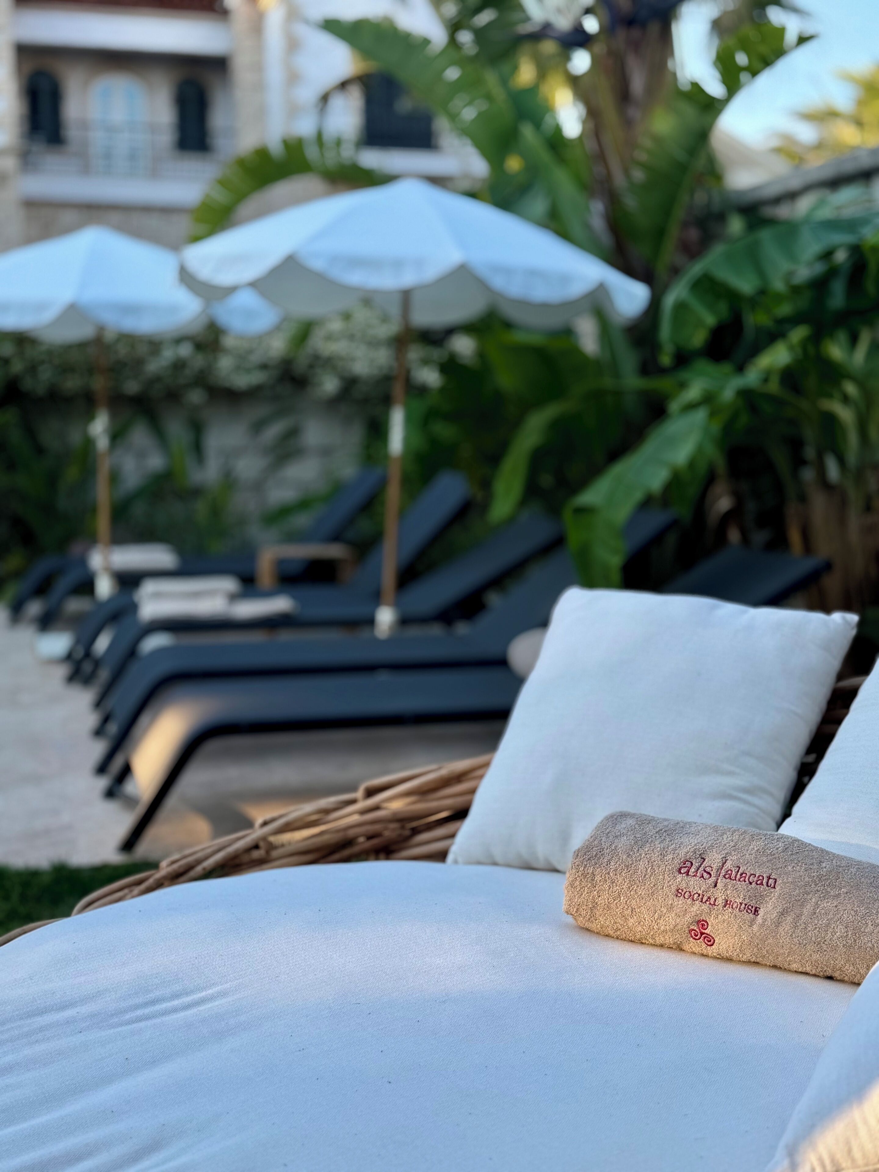 A row of sun loungers with white cushions and a towel with 'als.i.alecati SOCIAL HOUSE' text.