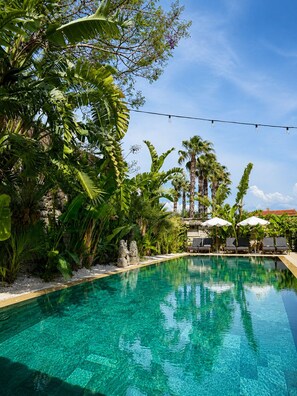 A clear pool surrounded by lush tropical plants and palm trees, with lounge chairs and umbrellas.