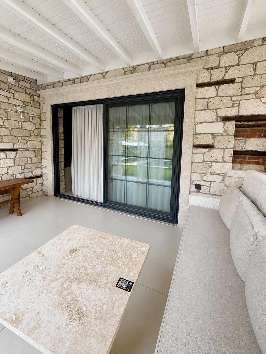 Una habitación con pared de piedra, puerta de vidrio corredera, sofá color beige y banca de madera.