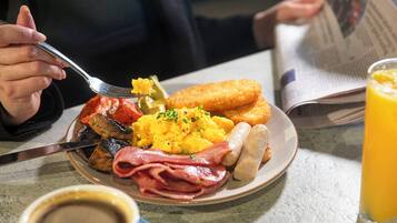 Une assiette de petit-déjeuner comprenant des œufs brouillés, du bacon, des saucisses, des pommes de terre rissolées et des toasts, accompagnée d’un verre de jus d’orange et d’une tasse de café.