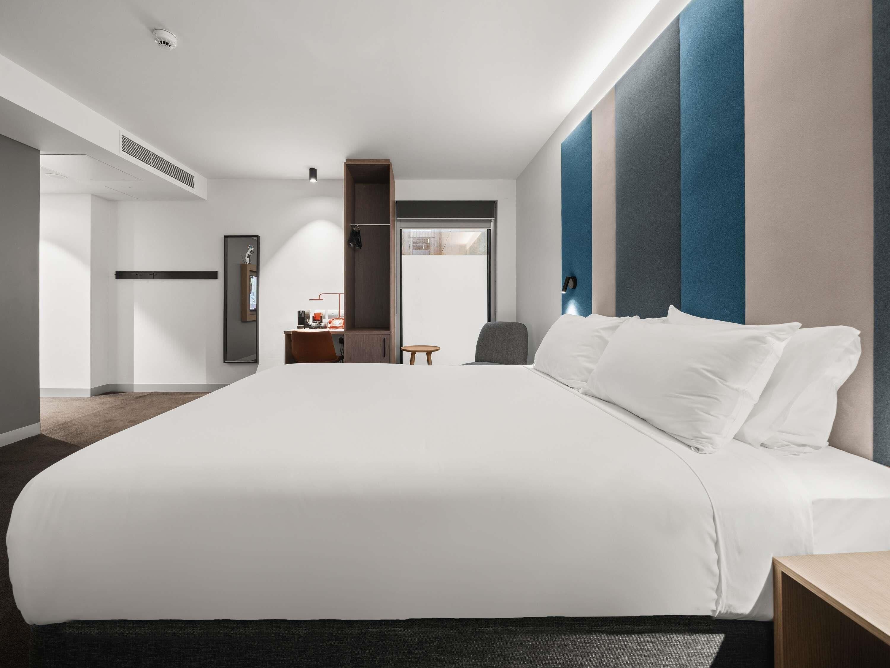 Ein modernes Hotelzimmer mit einem großen Bett, einem Schreibtisch, einem Stuhl, einem Kleiderschrank und einem Spiegel.