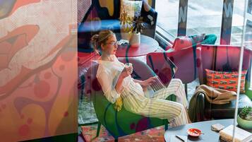 Un intérieur moderne où l’on voit une personne utilisant un ordinateur portable assise sur une chaise verte, une petite table avec des livres et un bol, et un mur coloré orné d’œuvres d’art abstraites.