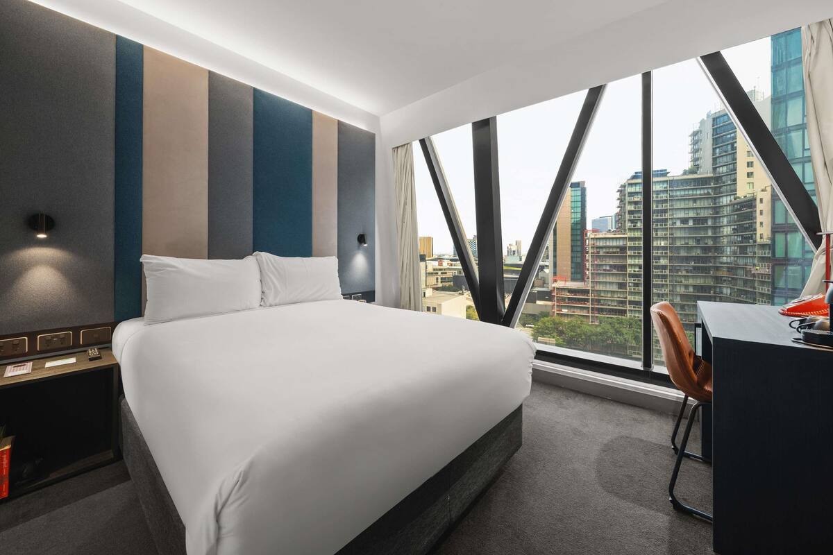 Ett modernt hotellrum med en stor säng, ett skrivbord och utsikt över stadsbilden genom stora fönster.