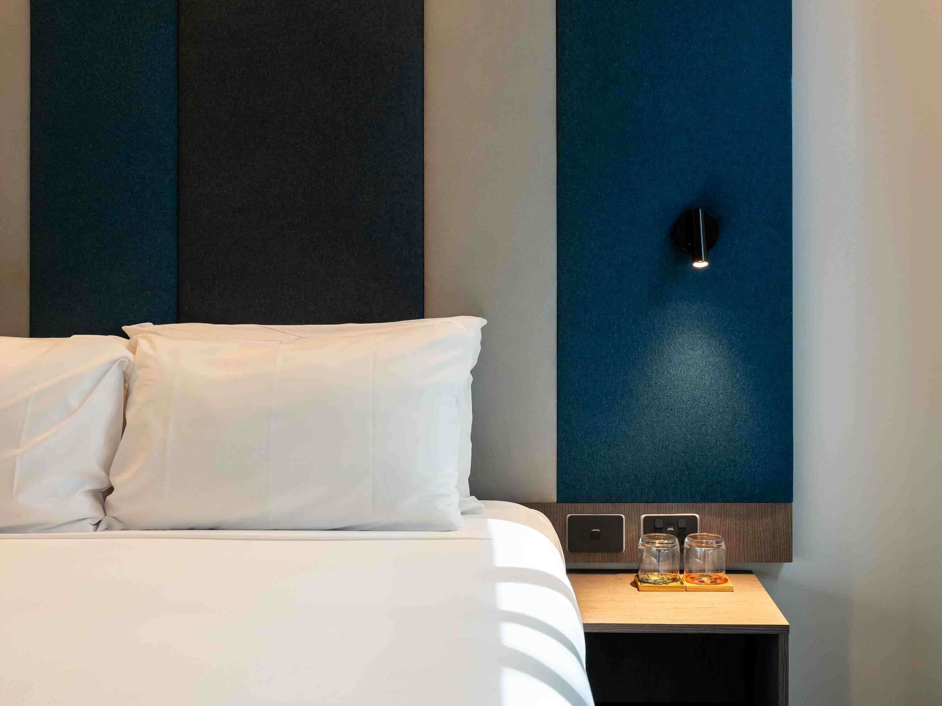 A hotel room with a bed, pillows, a nightstand with two glasses, and a wall-mounted light.