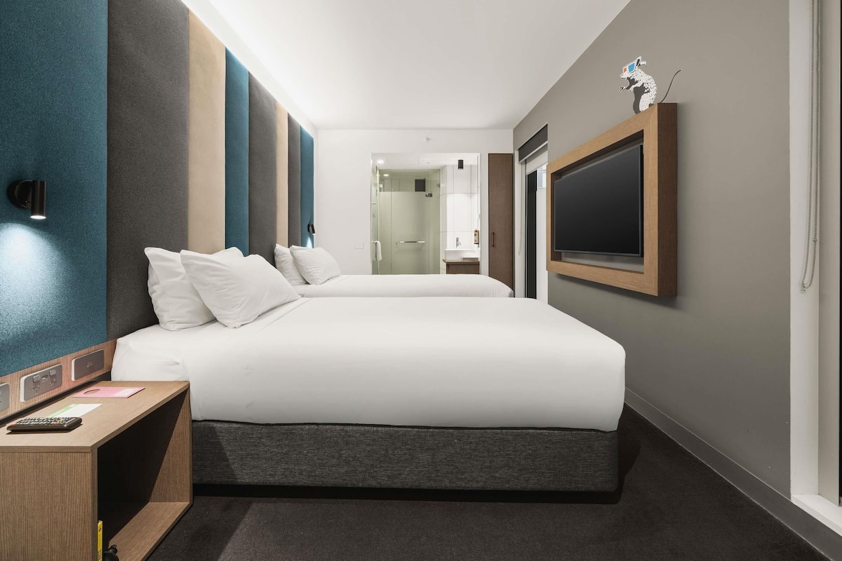 Ett modernt hotellrum med en stor säng, ett skrivbord med en dator, en tv och ett badrum som syns genom en glasdörr.