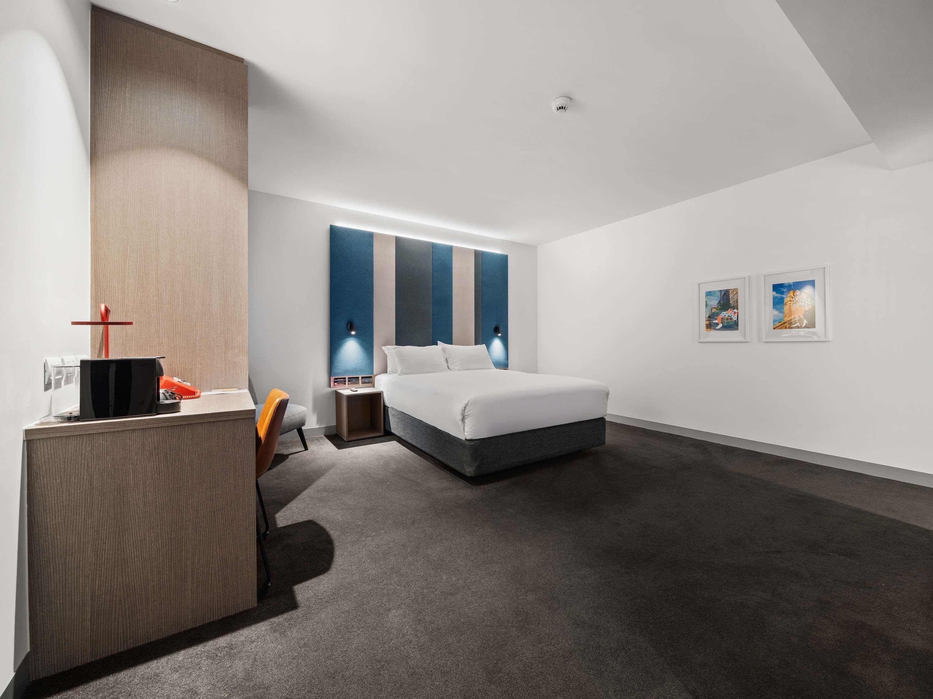 A modern hotel room with a bed, desk, chair, and artwork on the walls.