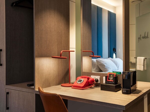 A hotel room with a wooden desk, a red rotary phone, a Breville coffee maker, and a mirror reflecting a bed with white linens.
