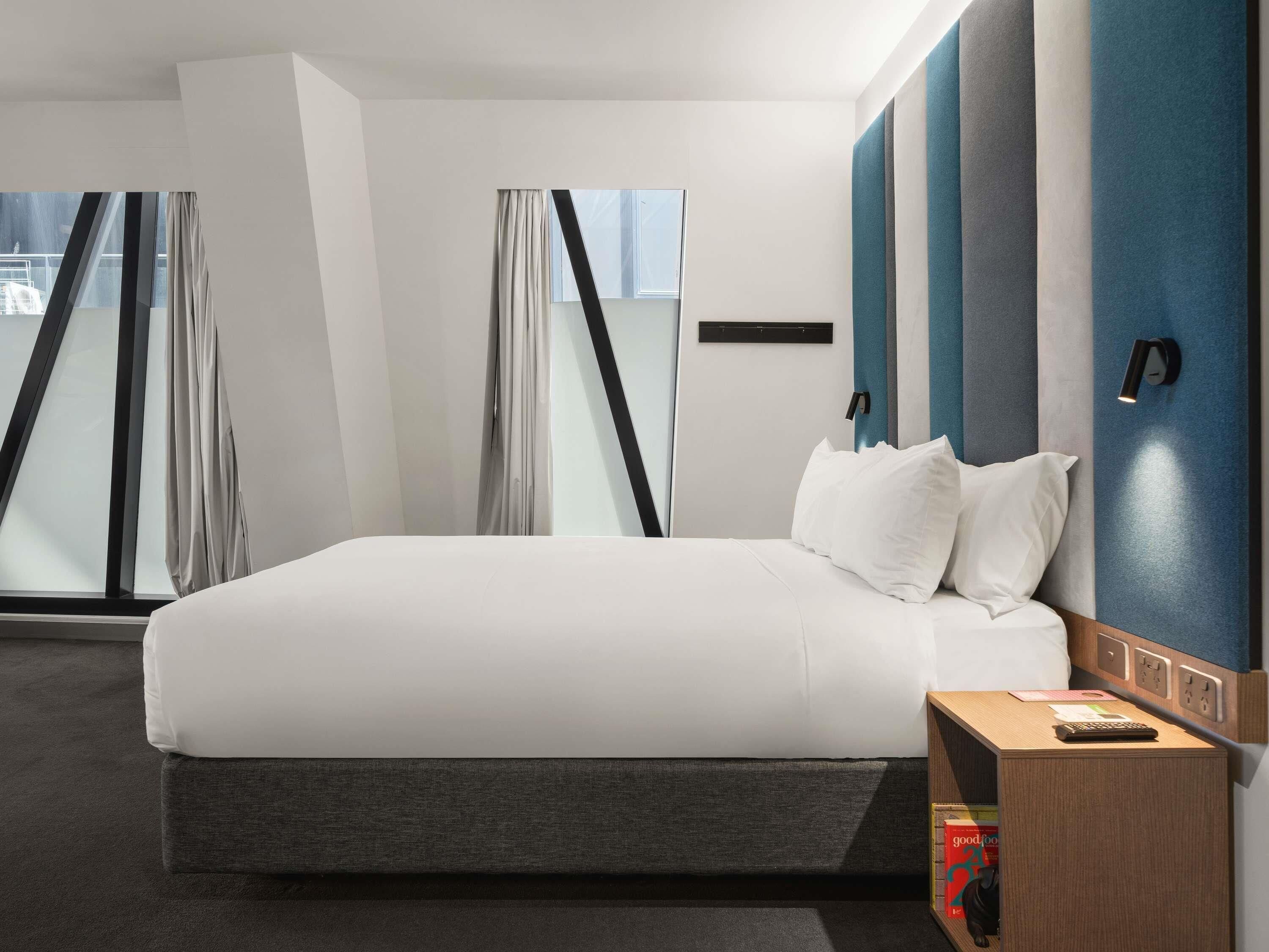 Une chambre d’hôtel moderne avec un grand lit, une table de chevet, une lampe fixée au mur et une fenêtre avec des rideaux.