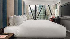 A modern hotel room with a large bed, a desk with a lamp, a TV, and a view of the city through large windows.