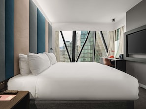 Una moderna camera d'albergo con un letto grande, una scrivania con una lampada, una televisione e vista sulla città da ampie finestre.