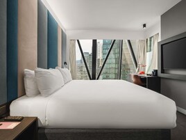Habitación de hotel moderna con una cama grande, un escritorio con lámpara, televisión y vistas a la ciudad a través de amplios ventanales.
