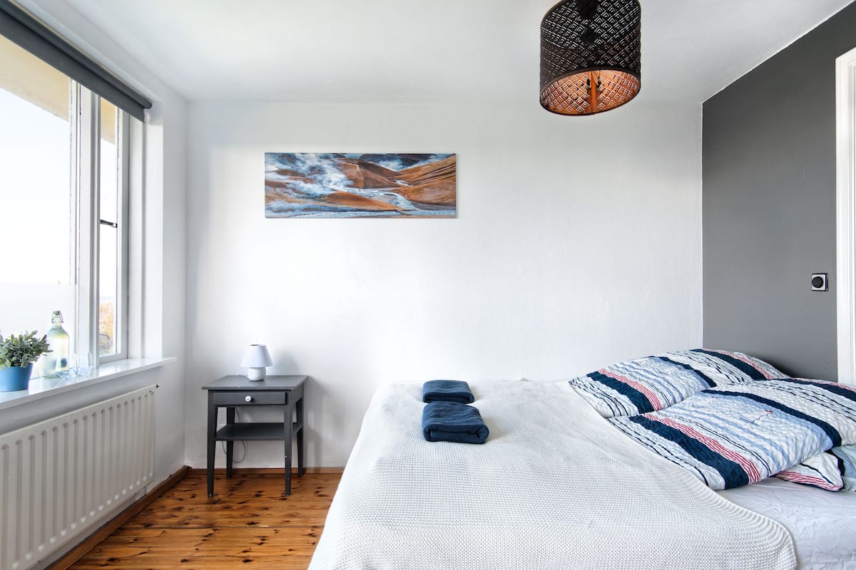A bedroom with a large bed, a small table with a lamp, a potted plant, and a painting on the wall.