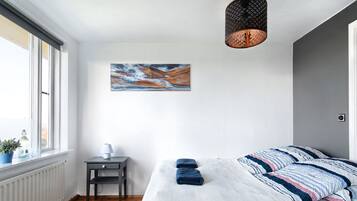 A bedroom with a large bed, a small table with a lamp, a potted plant, and a painting on the wall.