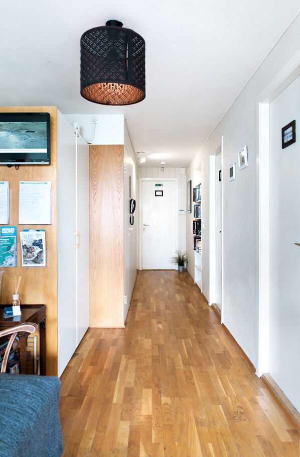Un pasillo con piso de madera, una lámpara colgante negra, una puerta de madera y una pared blanca con cuadros enmarcados y un televisor montado.